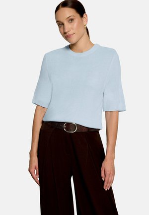 Kvinde med mørkt, trukket hår iført lyseblå kortærmet sweater, mørke bukser med vide ben og et brunt bælte med oval spænde.