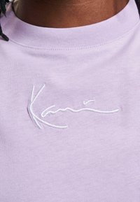 Šviesiai violetinė marškinė su apykakle, ant krūtinės išsiuvinėtas balty tekstas "Kani". Atrodo, kad medžiaga yra švelni ir lygi.