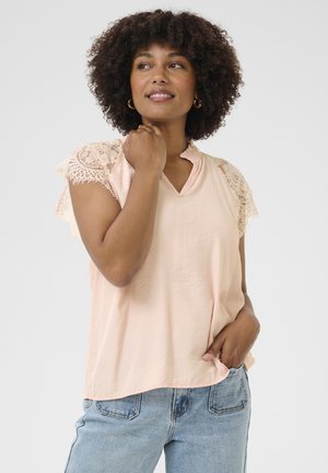 Blouse pêche clair avec des manches en dentelle, texture douce, design en V et coupe décontractée, portée avec un jean bleu clair.