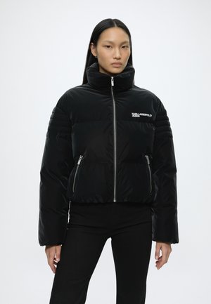 Vrouw draagt een zwarte donsjas met hoge kraag, ritssluiting aan de voorkant, ritssluitingzakken en het logo "Karl Lagerfeld Jeans" op de borst.