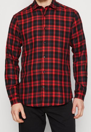 Hombre con una camisa de botones de manga larga a cuadros rojos y negros y pantalones negros, de pie frente a un fondo liso.