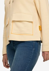 Chaqueta cremoso con cremallera, dos bolsillos delanteros, costuras naranjas contrastantes y una etiqueta lateral con un detalle del logotipo en naranja.