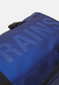 Blå vanntett ryggsekk med store pregete bokstaver, jevn tekstur, svarte stropper og klipslukking på toppen.