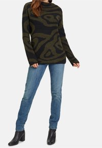 Femme portant un pull à motifs noir et vert olive, un jean bleu clair et des bottines noires, sur un fond uni.