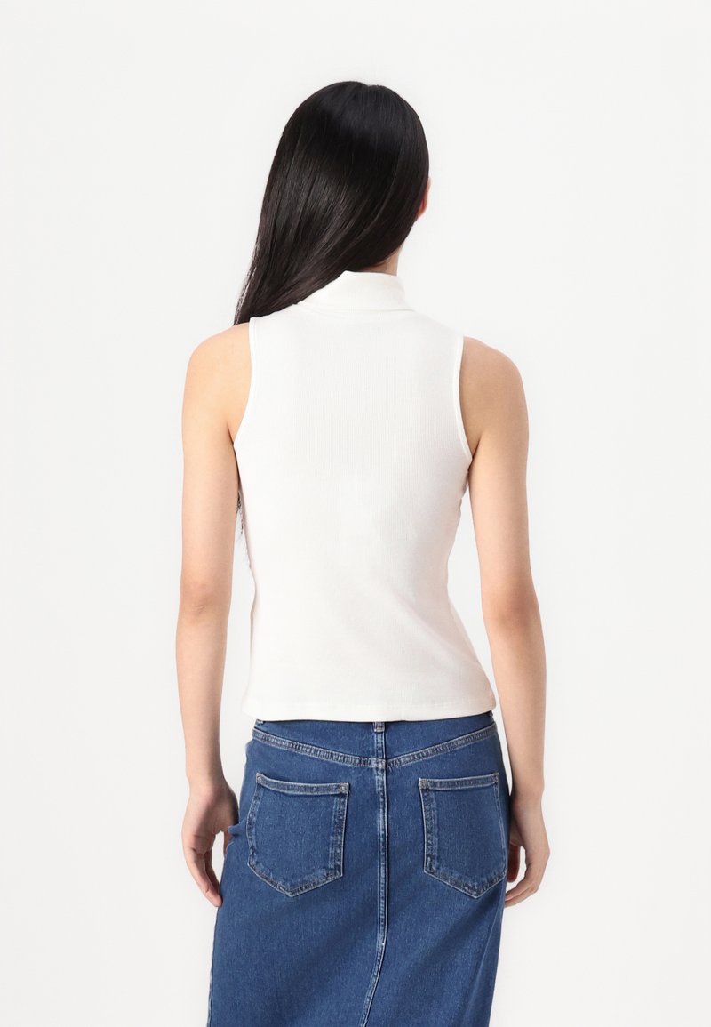 Top bianco senza maniche con collo alto e texture a costine, abbinato a una gonna di jeans blu con tasche posteriori e una silhouette aderente.