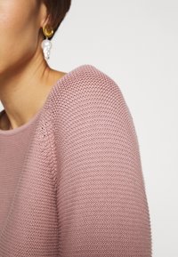 Gros plan d'une personne portant un pull en maille texturée rose et une boucle d'oreille en or avec un détail de perle blanche pendante.
