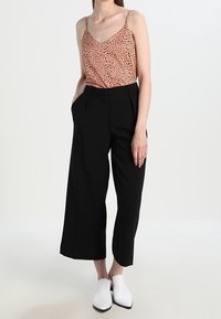 Pantalones de pierna ancha negros con pliegues frontales combinados con una camisola roja con estampado. La modelo lleva zapatillas blancas sin cordones. Fondo sencillo.