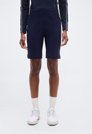 Persoon die marineblauwe knielange short, witte sokken, witte sportschoenen en een marineblauwe longsleeve met witte strepen op de mouwen draagt.