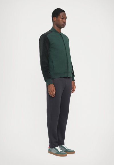 Grön bomberjacka med svarta ärmar, kombinerad med mörkgrå sweatpants och gröna sneakers med texturerade detaljer. Minimalistisk design.