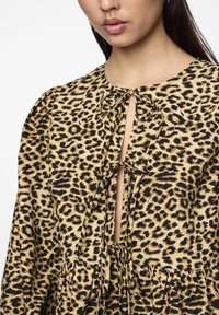 Blusa con stampa leopardo in beige e nero, con un'apertura a occhiello, dettaglio con nodo al collo e texture arricciata nella parte inferiore.