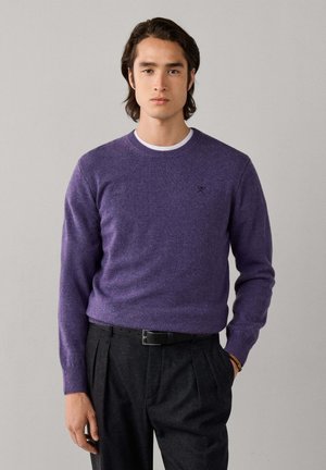 Joven con un suéter morado sobre una camisa blanca, pantalones oscuros plisados con cinturón negro, de pie frente a un fondo claro y liso.