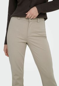 Pantalon beige à la texture lisse, doté d'une coupe ajustée et de poches latérales. Associé à un cardigan marron foncé.