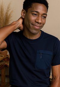 Marineblauwe T-shirt met korte mouwen en een textuurzak met een ritssluiting. Soepele stof met een ontspannen pasvorm, model weergegeven in een neutrale omgeving.