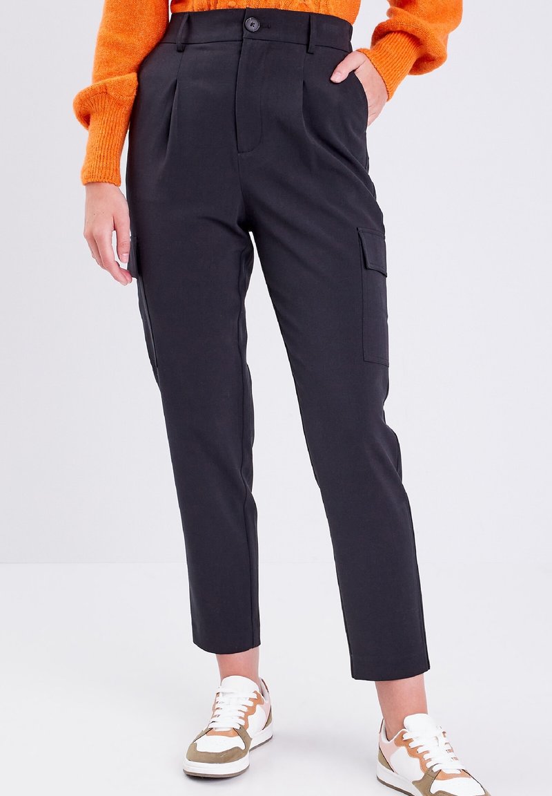 Person wearing black high-waist cargo pants with side pockets, orange sweater, and white sneakers with beige accents standing against a white background.