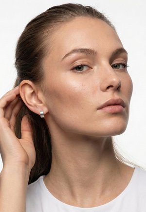Jeune femme à la peau claire et aux cheveux tirés en arrière ajustant une boucle d'oreille argentée en forme d'anneau, portant un haut blanc, expression neutre, fond blanc.