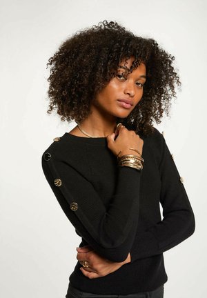 Maglione nero lavorato a maglia con texture a costine, scollatura rotonda e dettagli in bottone color oro sulle maniche. Modello che indossa diversi braccialetti.