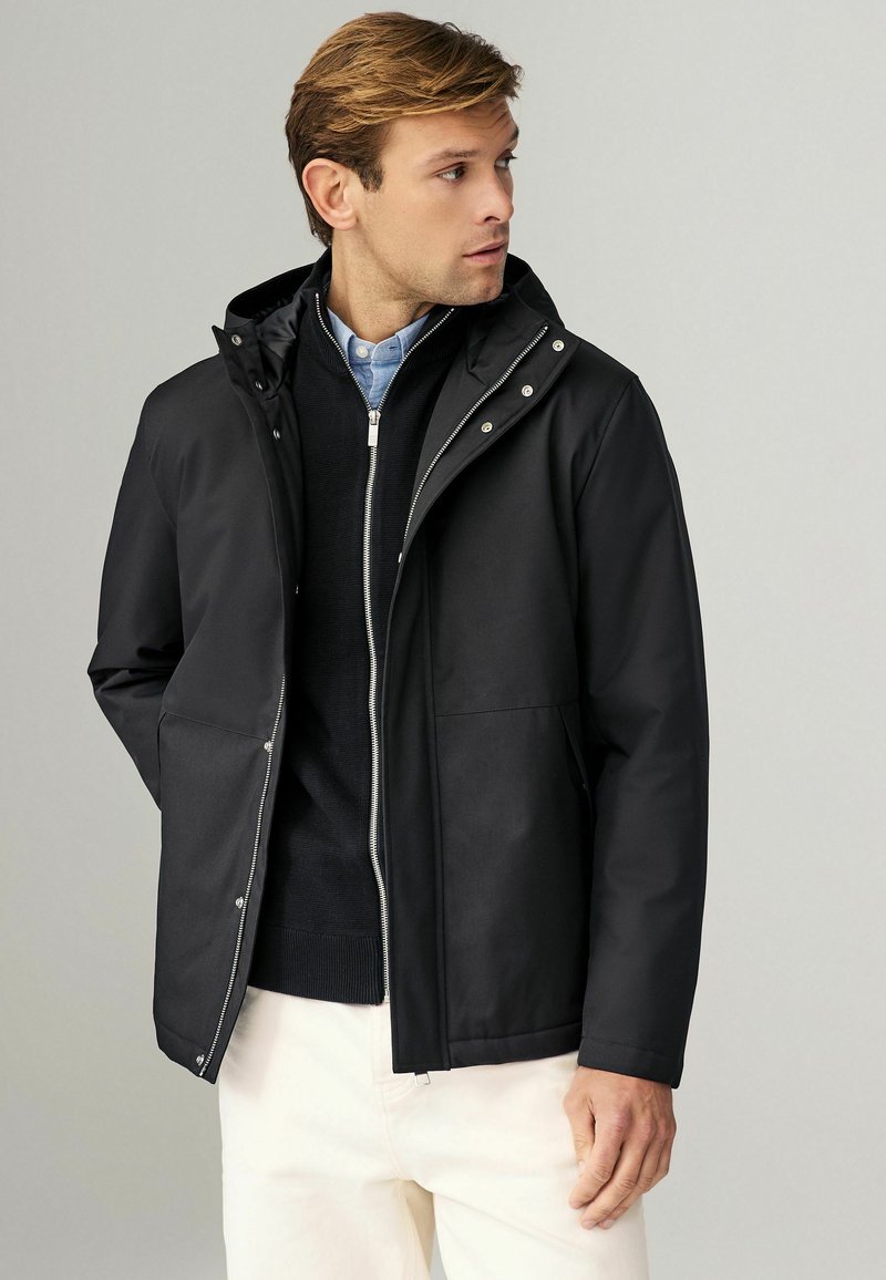 Giacca impermeabile nera con cappuccio, hardware argentato, due tasche frontali e chiusura con zip. Indossata sopra un maglione scuro e pantaloni chiari.