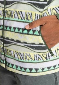 Chaqueta de forro polar con un bolsillo con cremallera, con un diseño estampado en verde, amarillo, gris y blanco. La tela texturizada mejora la calidez.