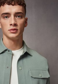 Camisa de botones verde claro con cuello, que presenta un bolsillo en el pecho y botones a presión, usada sobre una camiseta blanca de cuello redondo.