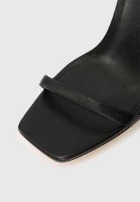 Μαύρο σανδάλι με ψηλό τακούνι, με φαρδιά, επίπεδη τετράγωνη μύτη και λεπτό λουράκι πάνω από το πόδι, κατασκευασμένο από λείο δέρμα με ματ τελείωμα.