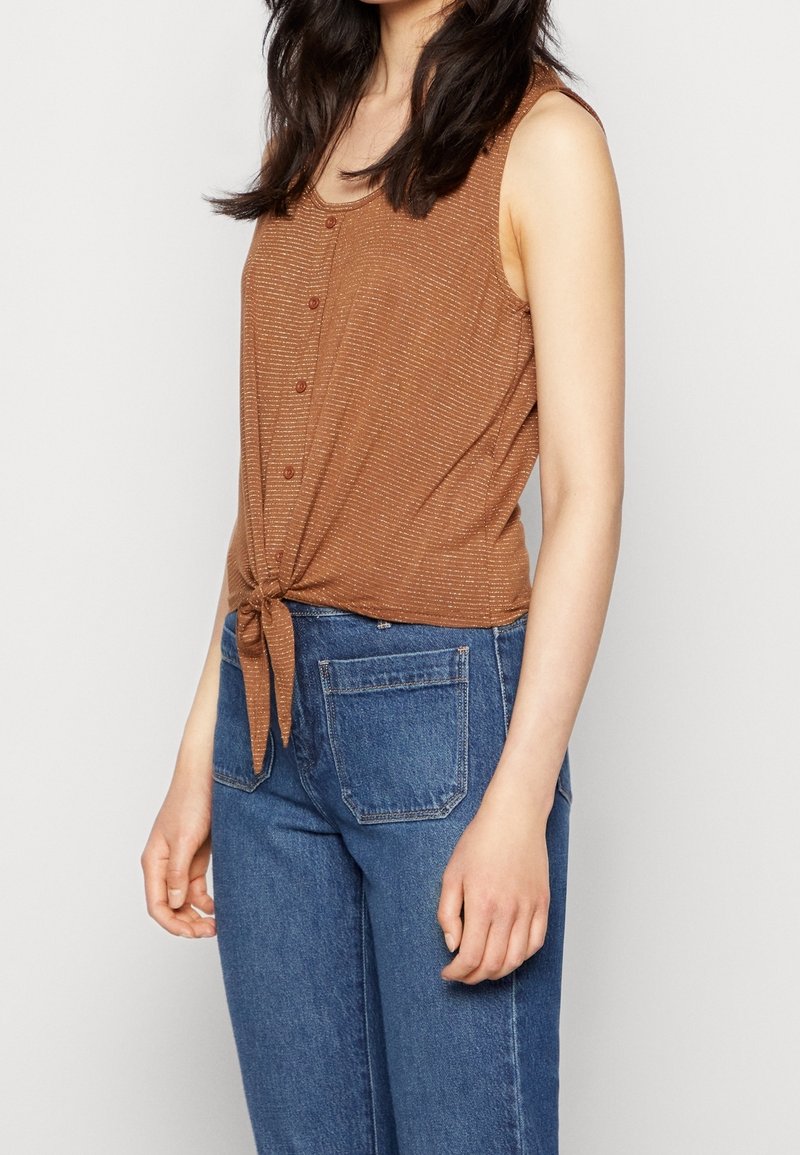 Bruine mouwloze top met een strik in de taille, met een knopenfront en een subtiele gestreepte textuur, in combinatie met blauwe high-waisted jeans.