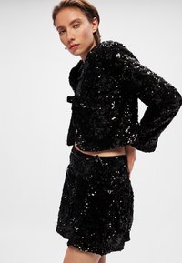 Svart paljettkoordset som innehåller en kort jacka med rosett och en minikjol med dragsko. Texturerad, glänsande yta med en figursydd design.