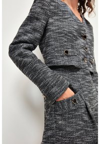 Chaqueta texturizada en blanco y negro con un diseño cropped, botones en tono dorado y dos bolsillos frontales. Presenta mangas largas y un escote en V.
