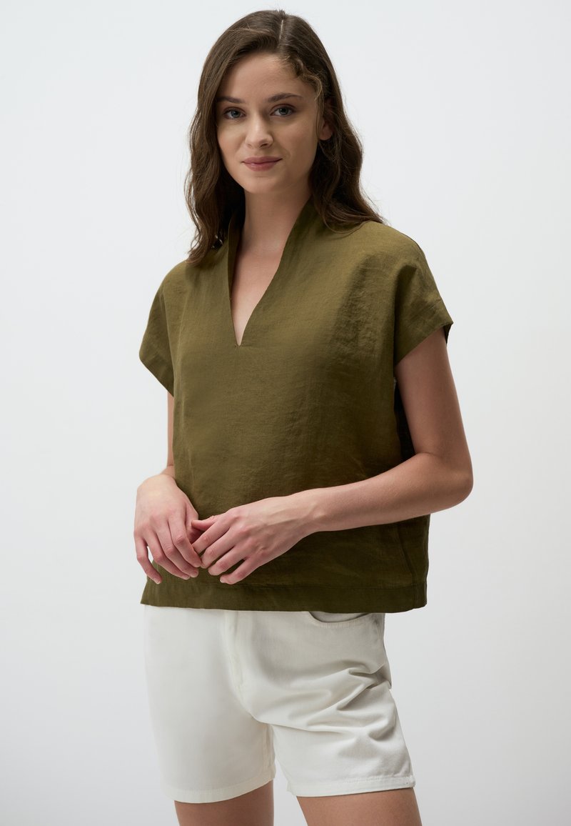 Top in lino verde oliva con scollatura a V, maniche corte e vestibilità ampia, abbinato a pantaloni corti bianchi. Il tessuto testurizzato crea un look casual.