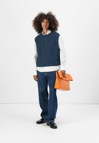 Navyblauer Strickwesten mit Zopfmuster, darunter ein weißes Hemd, blaue Jeans und eine orangefarbene Ledertasche. Schwarze Schuhe runden das Outfit ab.