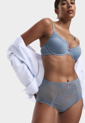 Vrouw in een blauwe kant beha en hooggesneden ondergoed, die een wijde witte gestreepte blouse aanpast tegen een effen achtergrond.