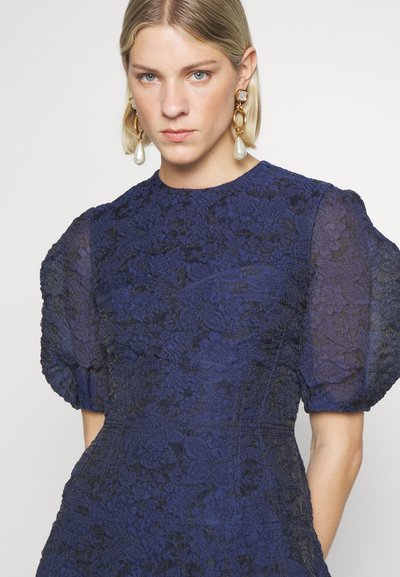 Vestido de renda azul-marinho com mangas bufantes transparentes, padrão floral texturizado, corpete justo e decote redondo. Grandes brincos de declaração.