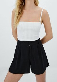 Top corto bianco a coste con spalline sottili abbinato a pantaloni corti neri a vita alta con pieghe. Texture lisce e design semplice.