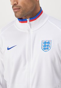 Biela bunda na zips z hladkej látky, s modrou logom Nike a erbom Anglicka so vzorom leva, obrysovaná červenými a modrými akcentmi.