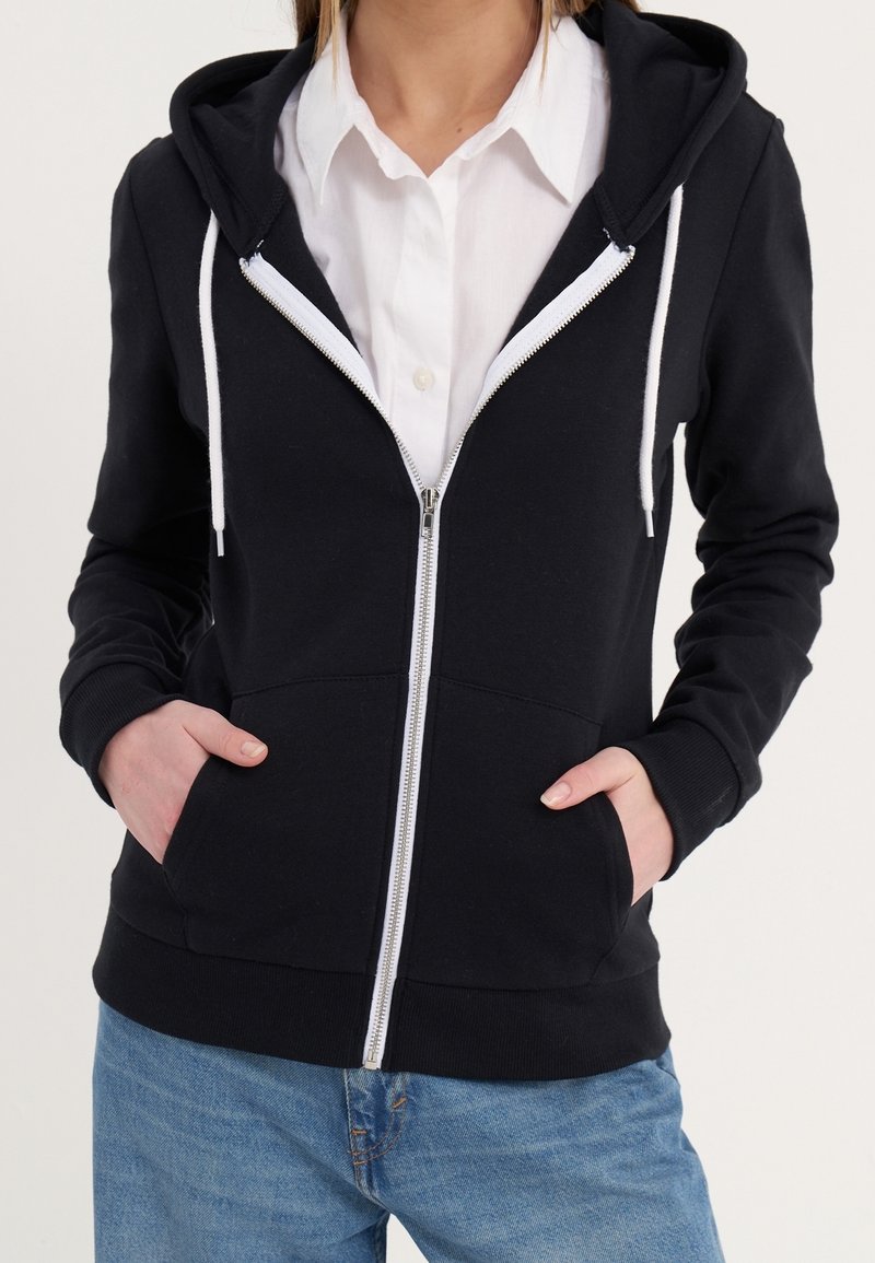 Personne portant un sweat à capuche noir zippé avec des cordons blancs par-dessus une chemise blanche à boutons et un jean bleu, mains dans les poches du sweat.
