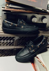 Černé kožené lodní boty Vans se zlatými průchodkami položené na a pod vintage audio přijímačem obklopeným vinylovými deskami.
