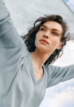 Kvinde med krøllet hår iført en lyseblå V-udskåret sweater, der løfter armene mod en delvist skyet himmel i baggrunden.