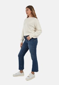 Maglione bianco a macchie con maniche lunghe abbinato a jeans blu in denim e sneakers bianche con dettagli dorati. Modella in piedi di lato.
