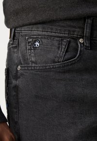 Jean en denim foncé à coupe slim, avec des détails de couture subtils, un logo bleu sur la poche avant et des accents en métal argenté.