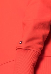 Červená mikina s ribovacími manžetami, se malým logem v námořnické, bílé a červené barvě na rukávu. Hladká textura a uvolněný střih.