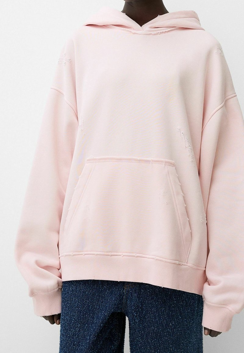 Osoba oblečená v nadměrné světle růžové mikině s přední kapsou a texturovaných modrých kalhotách, stojící na pozadí bez vzoru.