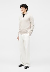 Mees seisab, kandes beeži neljandiku lukuga kampsunit, valgeid laiade säärega pükse ja musti kingi, käed taskutes ühevärvilise tausta ees.