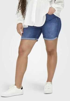 Denim shorts met een opgerolde zoom, in een donkerblauwe wassing, gedragen met een witte textuur blouse en witte sneakers.
