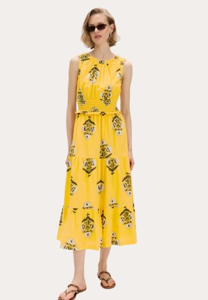 Kvinde iført gul blomsterprintet midi-kjole uden ærmer med lagdelt skørt, brune sandaler, solbriller og guld øreringe, stående mod en ensfarvet baggrund.