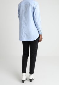 Camicia button-up azzurra con orlo curvo, maniche lunghe e yoke posteriore; abbinata a jeans skinny neri e stivaletti bianchi alla caviglia.