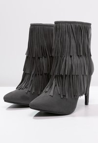 Bottes chelsea en daim noir avec bouts pointus et talons hauts, présentant de longs détails en frange le long du tige. Texture lisse et design élégant.