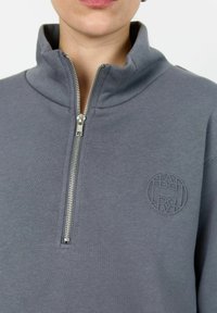 Siva zip-up dukserica z visokim ovratnikom, s srebrnim Zipom in okroglo vezeno logotipom na prsih. Mehek, pleten material.