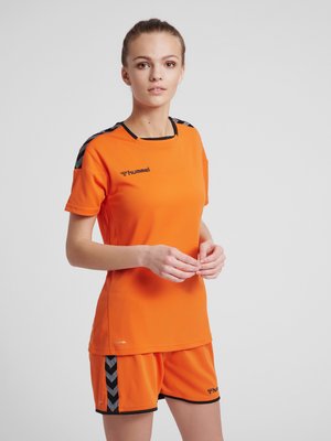 Ung kvinde står iført en orange sports trøje og shorts med sorte Chevron-striber på skuldrene og shortssiderne.