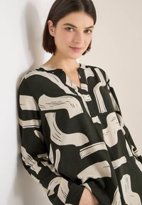 Bluzka z długim rękawem w ciemnozielonym materiale, zdobiona abstrakcyjnymi beżowymi wzorami przypominającymi pociągnięcia pędzla, z dekoltem w serek i zdobieniem na plisie.