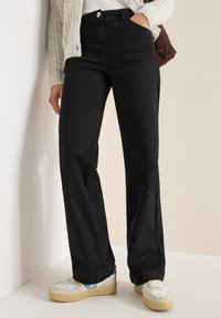 Jeans negros de pierna recta con bolsillos frontales y cintura alta, combinados con zapatillas blancas y crema. Tela de denim suave con una textura lisa.