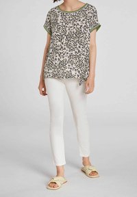Kortærmet top med hvid baggrund, sort og grøn leopardmønster, og en knude detaljeret ved kanten. Matcher med hvide stramme bukser og beige sandaler.